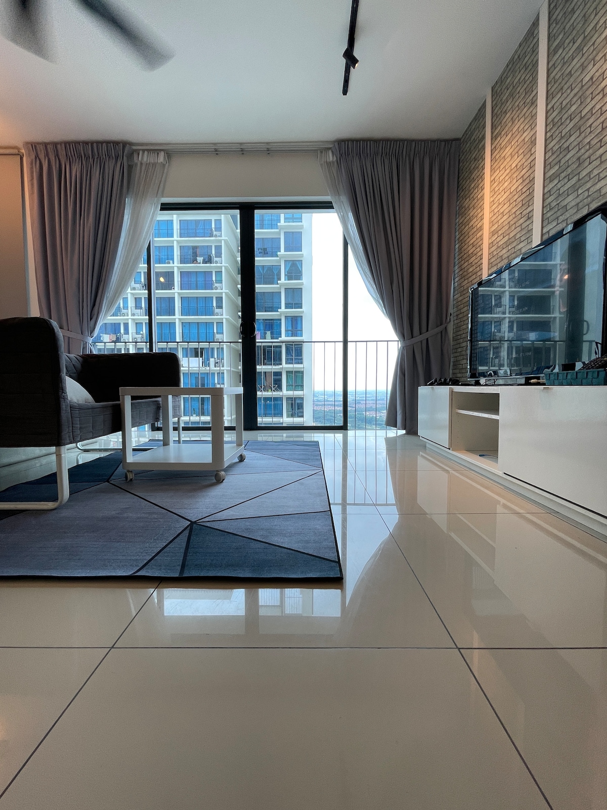 A spacious living area is highlighted by large windows, allowing natural light to fill the room. Modern furnishings include a comfortable seating arrangement and a sleek media unit with a TV. A patterned area rug adds texture to the tiled floor, creating a serene atmosphere.
