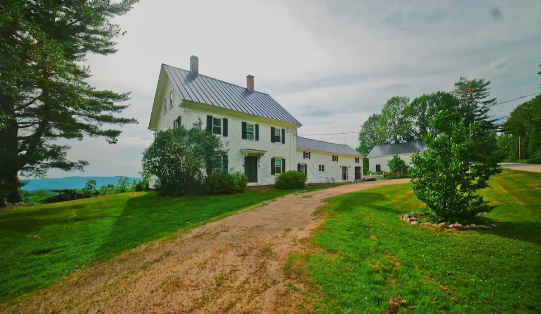 The historic farmhouse is set on a grassy expanse with a winding gravel driveway leading up to it. The white structure features black shutters and a metal roof, surrounded by trees and green landscaping, with a hint of distant mountains visible in the background.