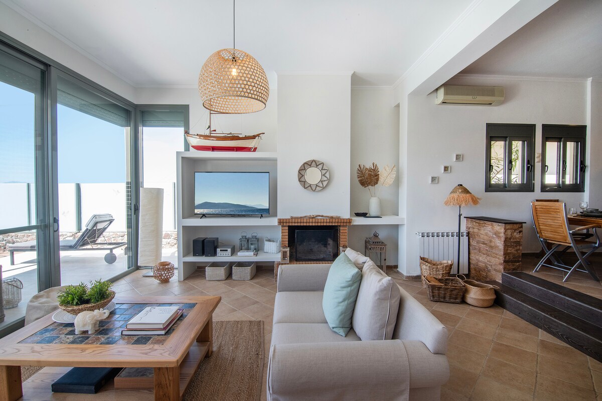 An open living space is highlighted by a light-colored sofa and natural textures. A wooden coffee table with decorative items is centered, while large glass doors allow natural light and provide access to the outdoor views. A flat-screen TV is mounted on the wall, adding a modern touch.