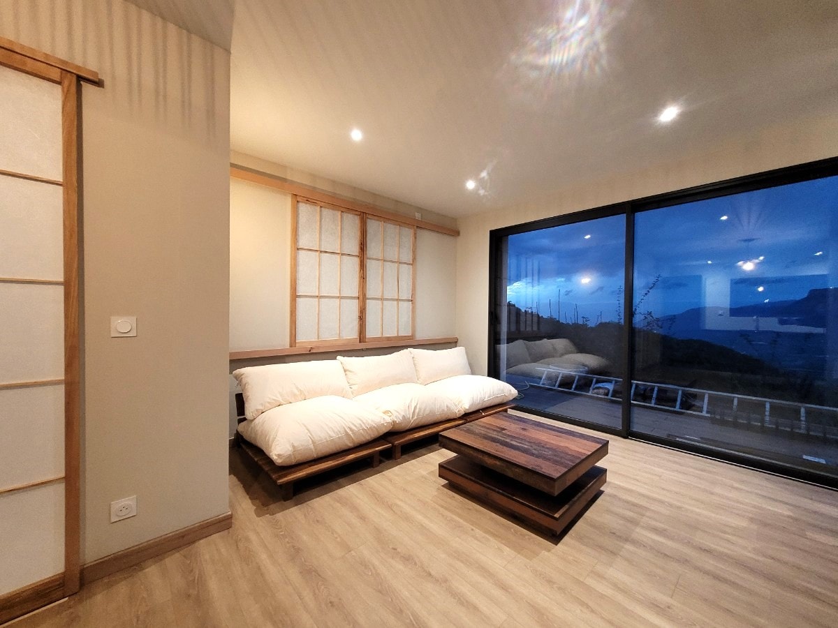 A spacious living area features a minimalist design with light wood flooring. A low wooden coffee table is positioned centrally, accompanied by a large, comfortable sofa with plush pillows. Expansive glass doors provide a scenic view of the outdoors and allow natural light to flood the room.