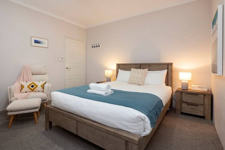 Ampia camera da letto con un letto matrimoniale con lenzuola morbide e un elegante copriletto blu. Include una comoda poltrona per il relax, comodini con illuminazione calda, ganci da parete per comodità e soffice pavimento in moquette.