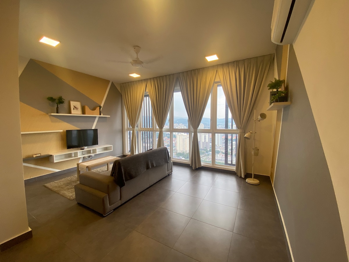 A modern living area is highlighted by large windows draped with soft, light-colored curtains. A grey sofa is positioned on a plush rug, facing a mounted television. Neutral tones and geometric accents create an open and spacious environment, complemented by a decorative plant.
