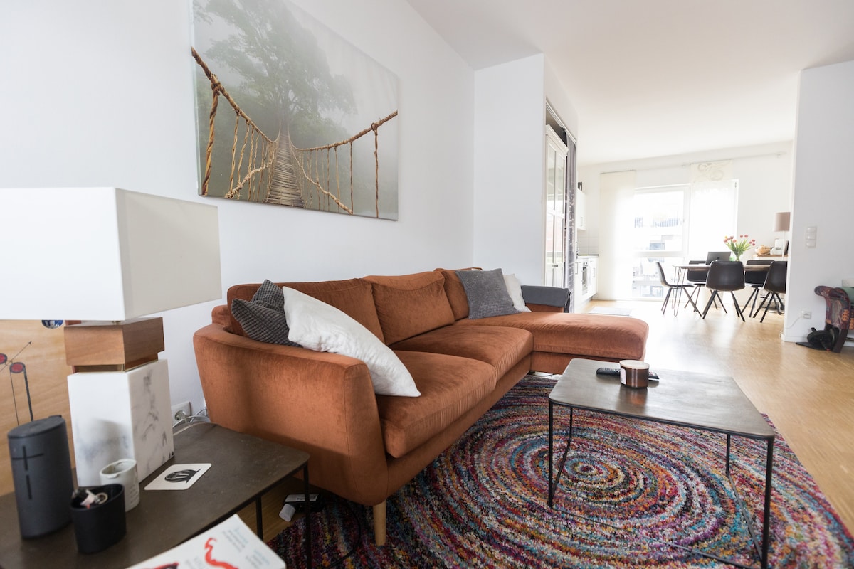 A spacious living area features an orange sectional sofa, complemented by a textured area rug. A coffee table rests in front, alongside decorative items. A large artwork of a bridge adorns the wall, while a dining space with a table and chairs is visible in the background.