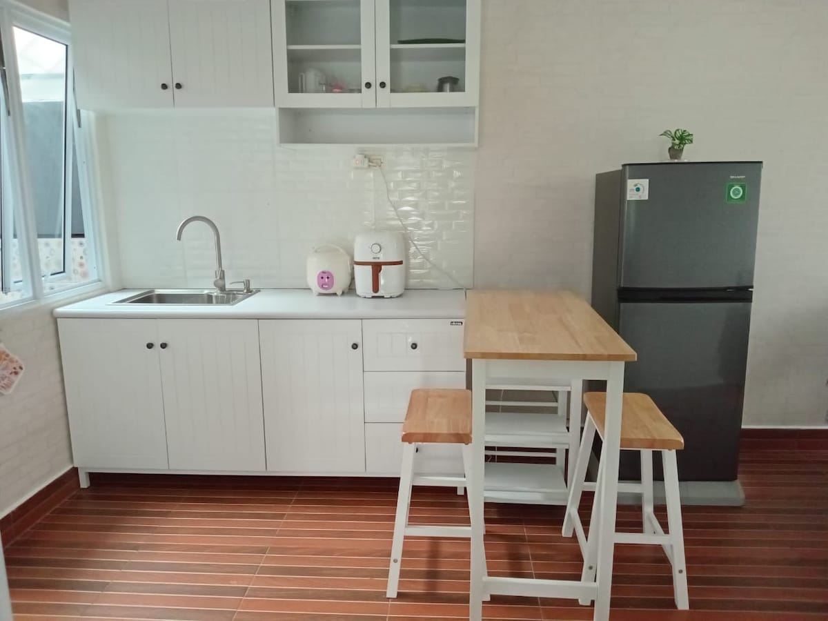 A modern kitchen space features white cabinetry and a stainless steel sink. A compact dining table with two wooden stools is positioned nearby. A refrigerator stands against the wall, and natural light flows in through the window, highlighting the clean design.
