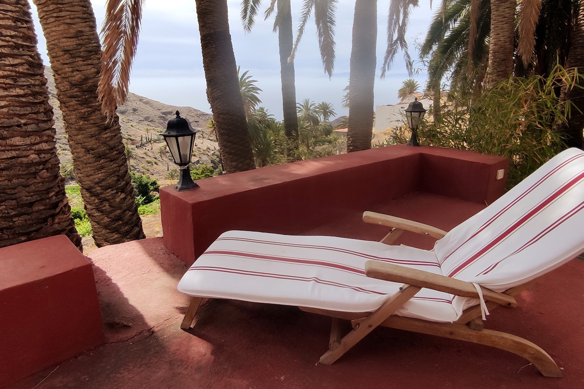 A shaded lounge area features a wooden recliner with white and red striped cushions. Surrounding palms create a serene environment, with a glimpse of distant hills and the sea visible in the background. Two lanterns are positioned nearby, enhancing the peaceful setting.