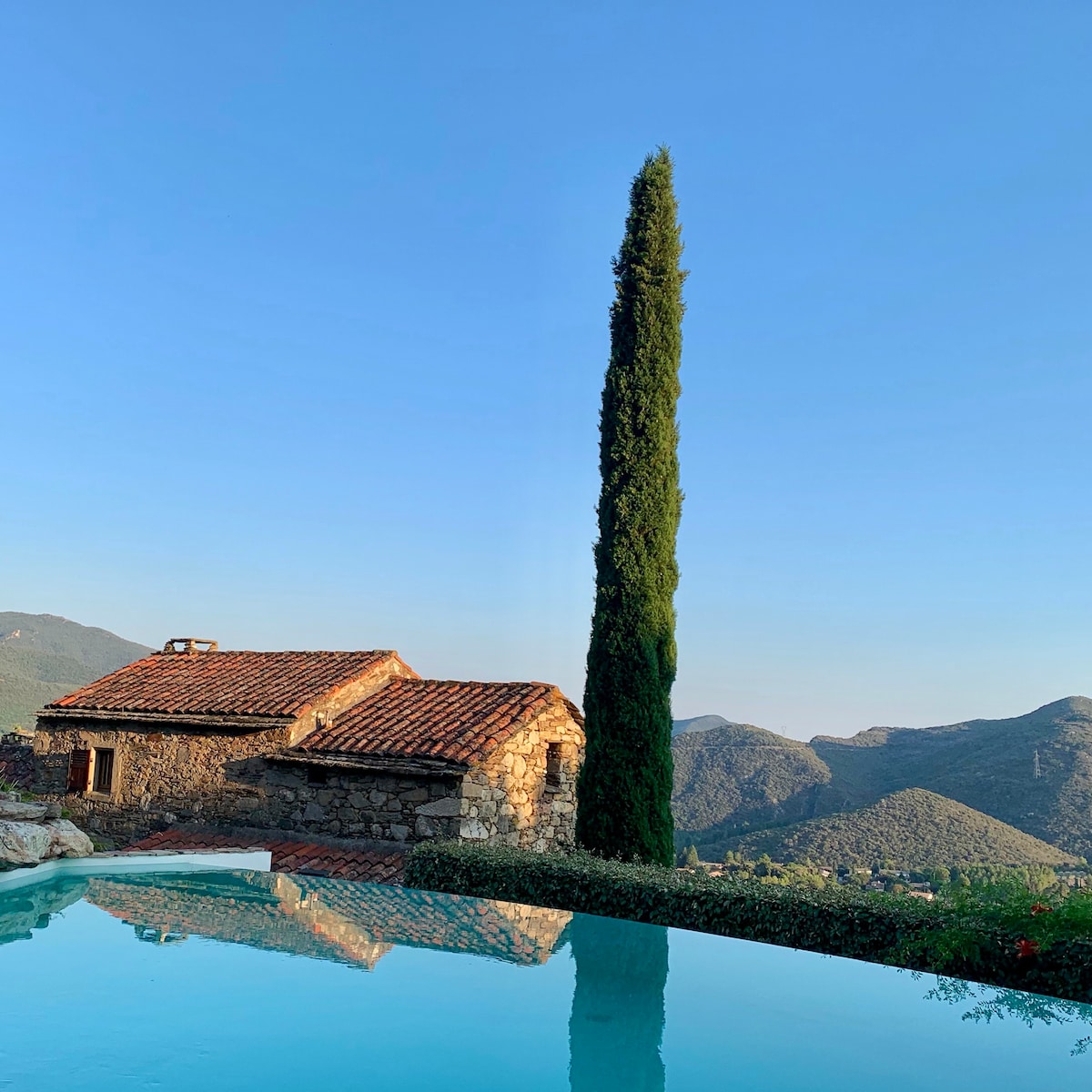 A stone building with a traditional tiled roof is visible near a calm pool, reflecting the surrounding landscape. A tall cypress tree stands prominently against a clear blue sky, framing the scenic view of rolling hills in the background.