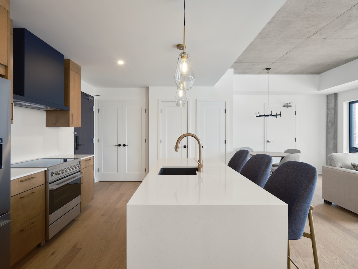A modern kitchen features sleek cabinetry in navy and wood tones, paired with stainless steel appliances. A large white countertop includes a sink, complemented by three upholstered bar stools. The open layout allows for natural light to illuminate the adjoining dining and living areas.