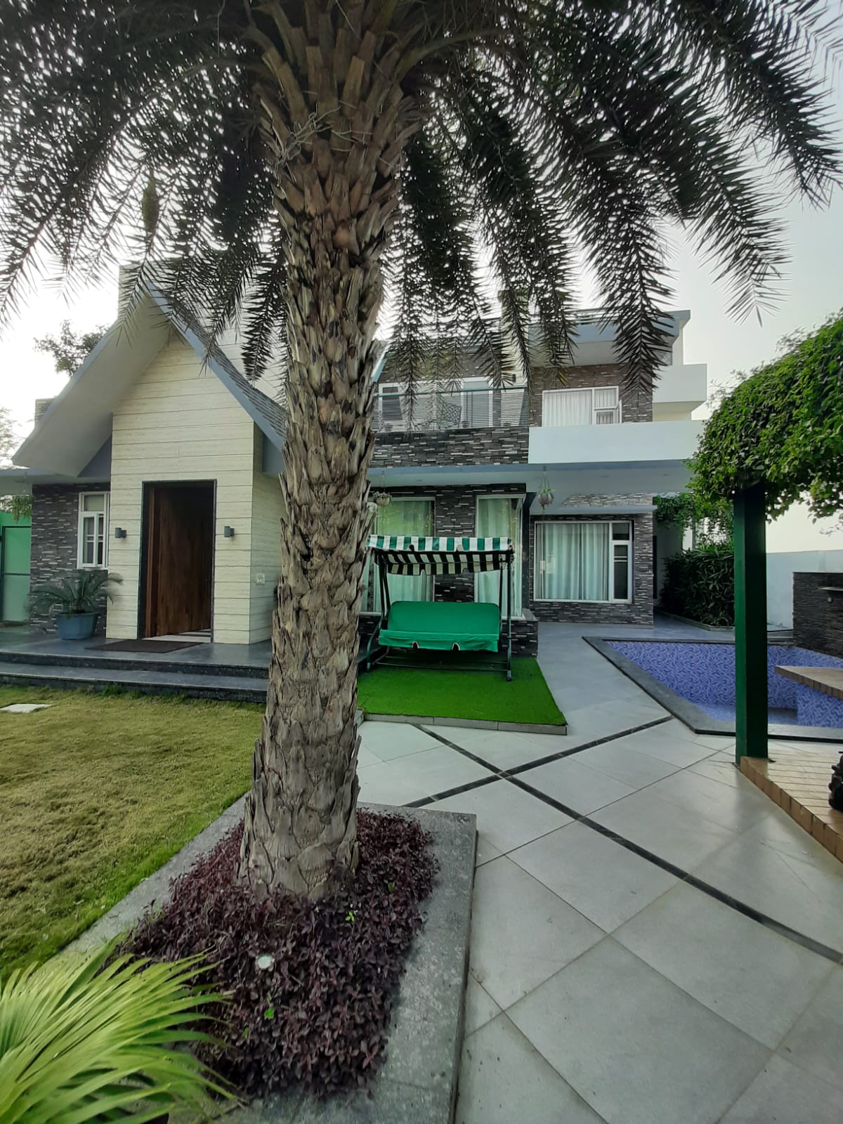 The villa exterior features a stone pathway leading to a light-colored wooden door framed by lush greenery. A vibrant palm tree stands prominently, while a relaxing swing sits on a manicured lawn, inviting relaxation in a serene setting.