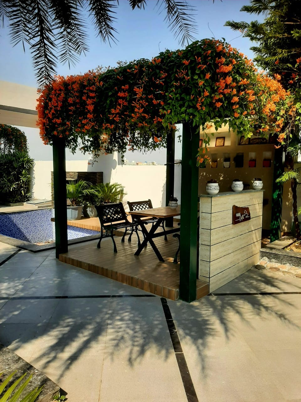 A dedicated outdoor seating area is framed by vibrant orange flowers, creating a serene space for relaxation. Two black chairs are positioned at a wooden table, with lush greenery and a glimpse of a pool visible in the background.
