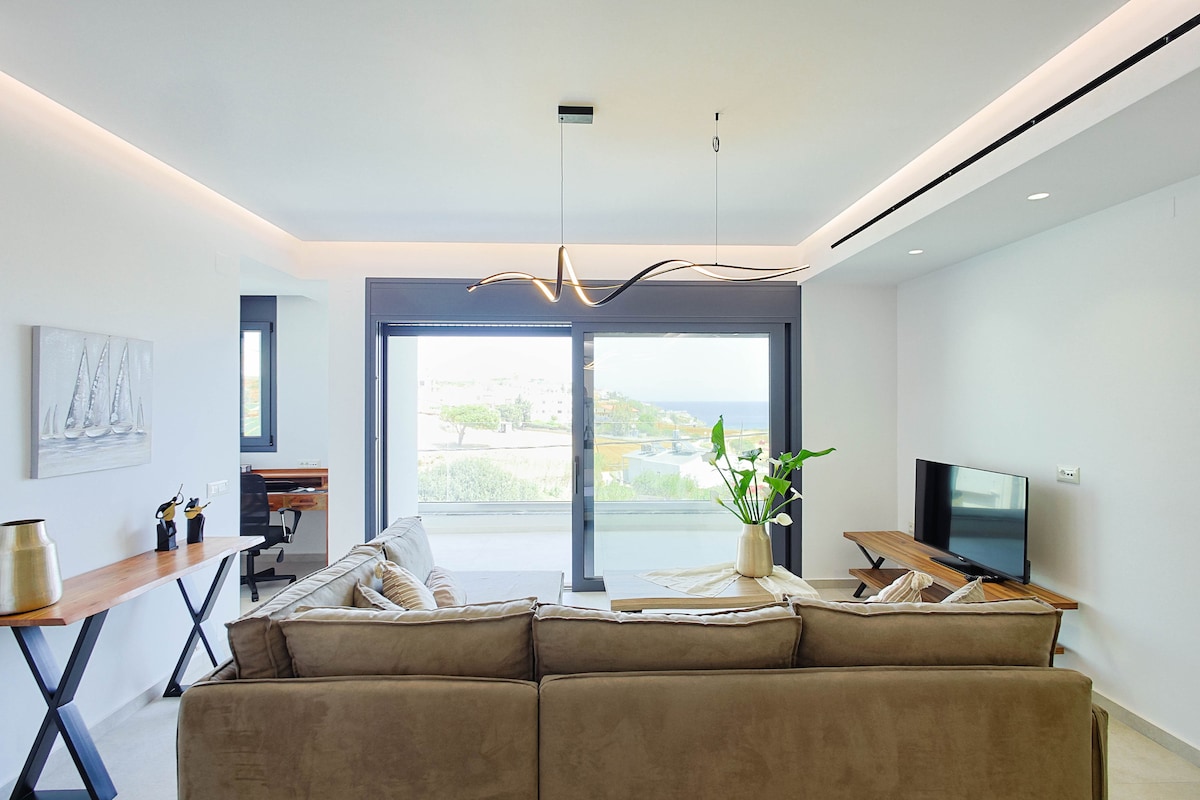 The living room features a modern corner sofa, a smart TV, and a coffee table. Large glass doors allow natural light to fill the space and provide access to a balcony with views of the surrounding area. A potted plant adds a touch of greenery.