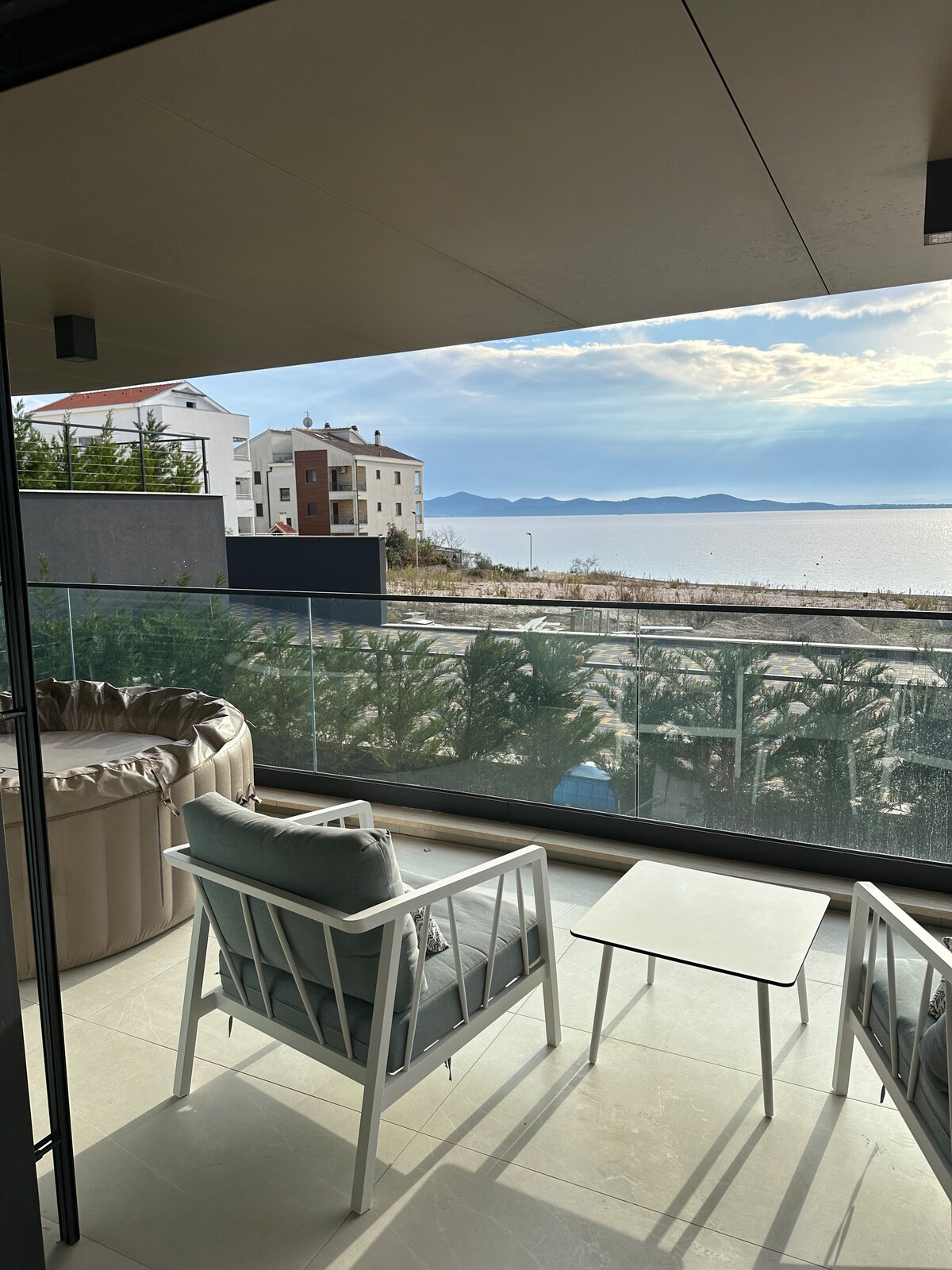 A private terrace features comfortable seating arrangements with a small table, overlooking the expansive views of the coastline and the shimmering sea. The surrounding area is adorned with greenery, providing a serene backdrop against the modern architectural elements of the property.