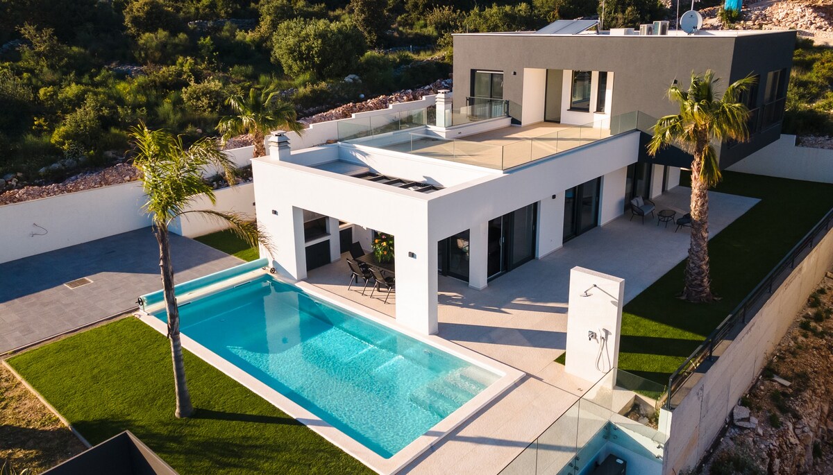 The villa showcases a modern architectural design with large windows providing ample natural light. A spacious outdoor area features a heated pool surrounded by a patio and sun loungers, complemented by lush greenery and palm trees. The roof terrace offers expansive views of the surroundings.