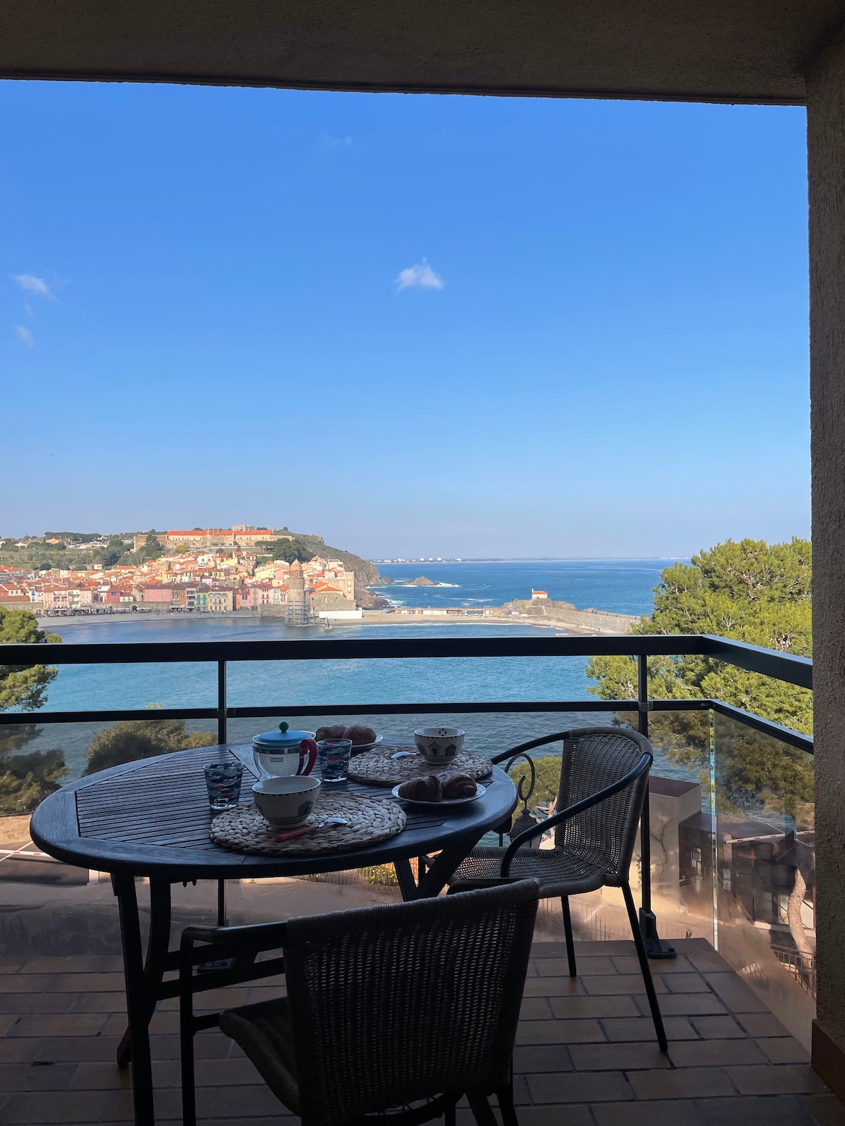 A balcony table is set for breakfast, featuring two cups, a small dish of pastries, and utensils. The view reveals the bay of Collioure, with vibrant buildings and coastlines under a clear sky. A serene atmosphere is provided by surrounding trees.