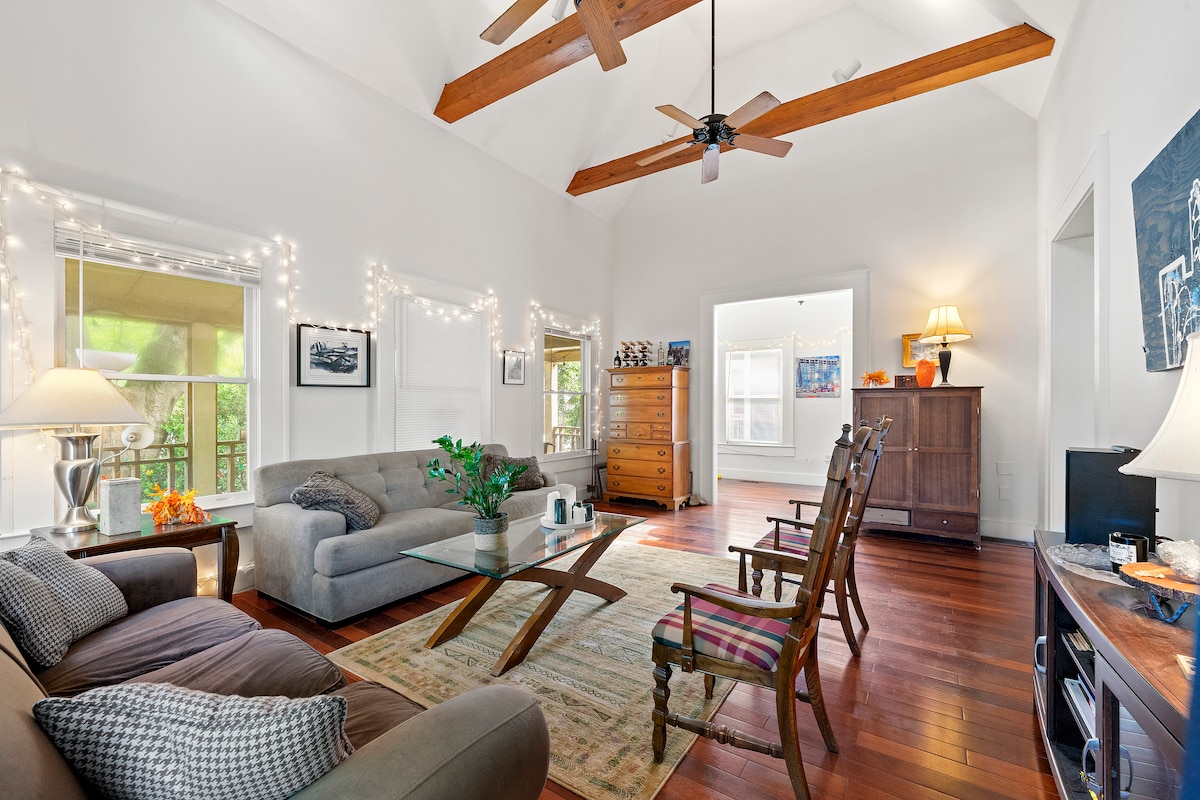 A spacious living area features tall ceilings with exposed beams. Soft seating is arranged around a geometric coffee table, while string lights softly illuminate the space. A vibrant area rug anchors the room, and large windows invite natural light, highlighting the warm hardwood floors.