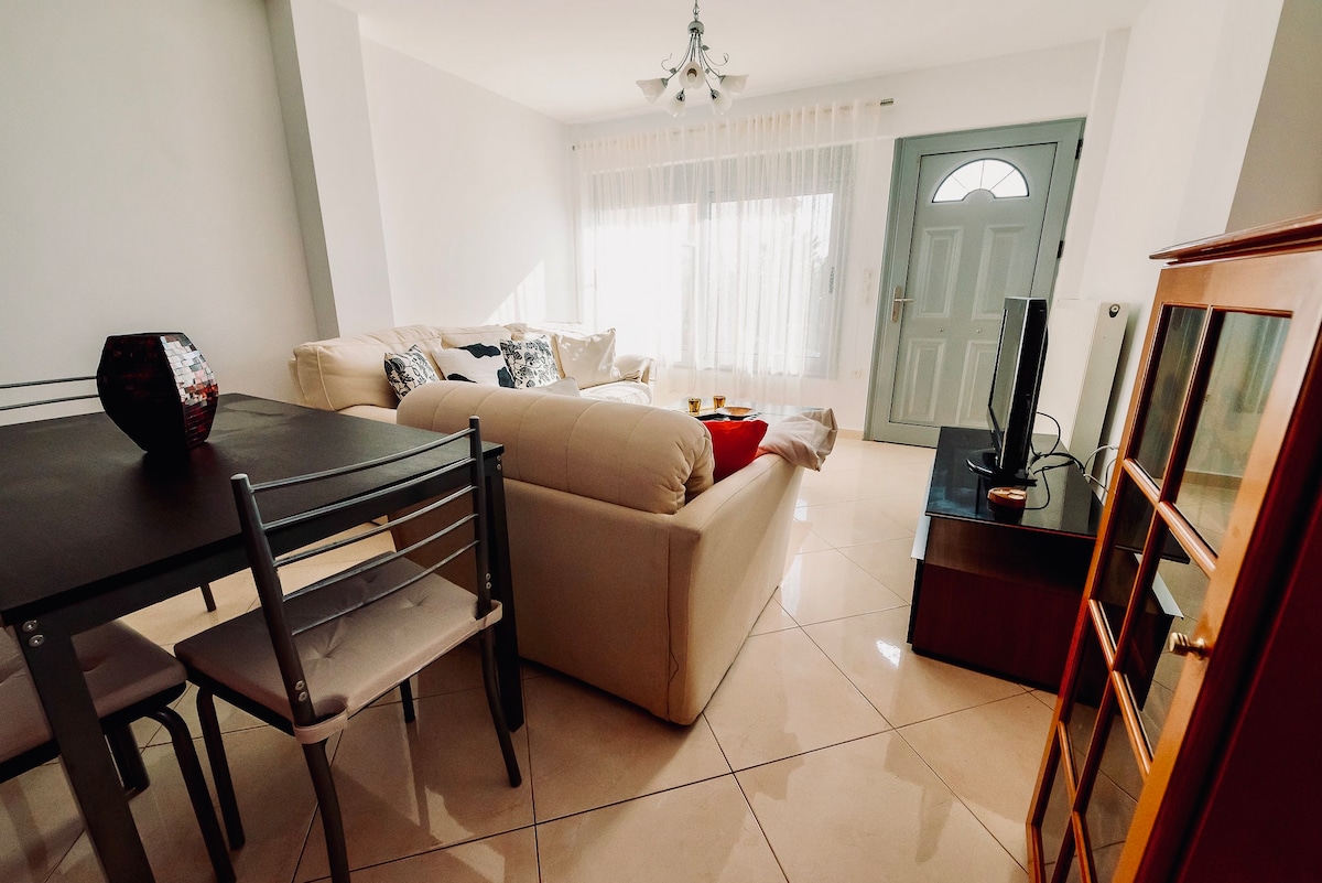 A bright living area displays a light-colored sectional sofa facing a TV stand. A wooden dining table with four chairs is positioned nearby. Natural light enters through large windows, accompanied by sheer white curtains and a door leading to the outside.