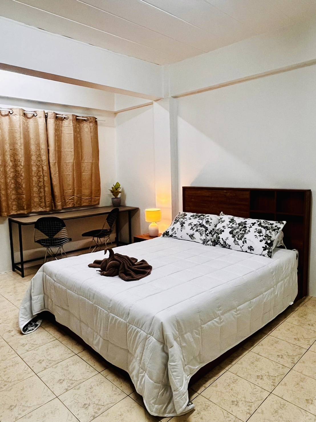 A serene bedroom features a spacious bed dressed in white linens with floral-patterned pillows. A small desk with two chairs is positioned nearby, complemented by a softly glowing lamp. Natural light enters through curtains, creating a calm environment.