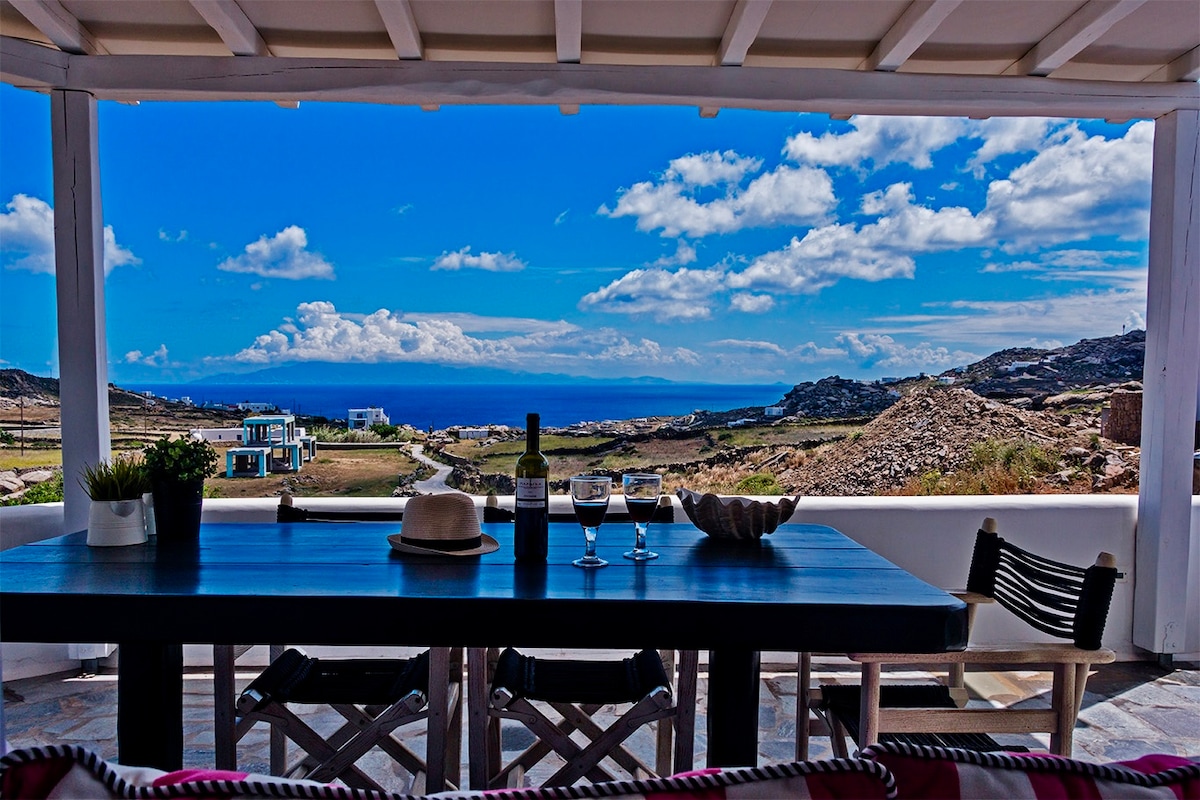 A dining area features a long wooden table set for four, with a bottle of wine and glasses positioned at the center. A wide view of the sea and surrounding landscape is visible through the open space, framed by a white pergola against a vibrant blue sky.