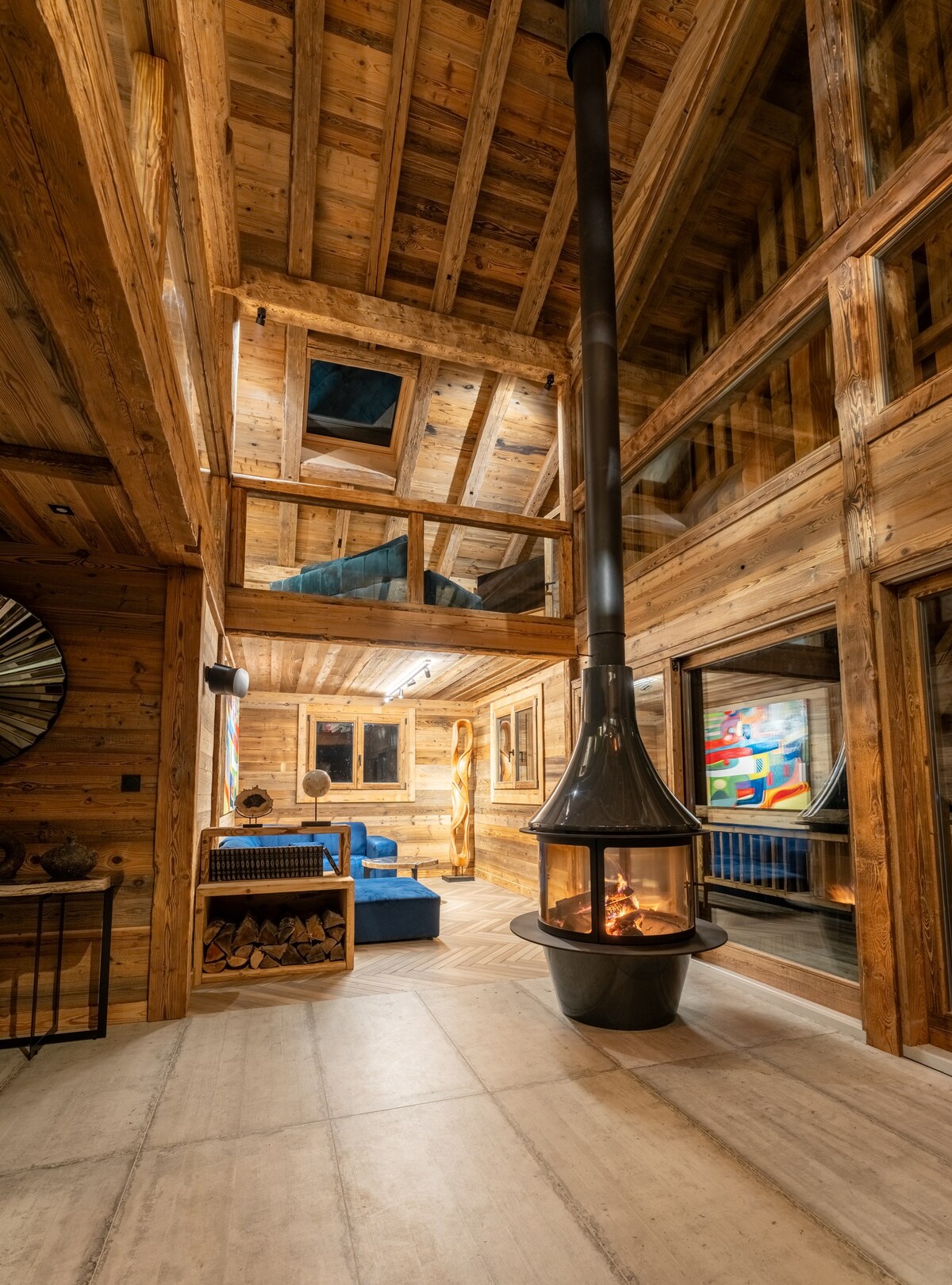 A central fireplace features a modern design and serves as a focal point in the cozy wooden interior. Large windows allow natural light to illuminate the space, while a comfortable seating area with blue upholstery is visible, complemented by natural wood accents and artwork in the background.