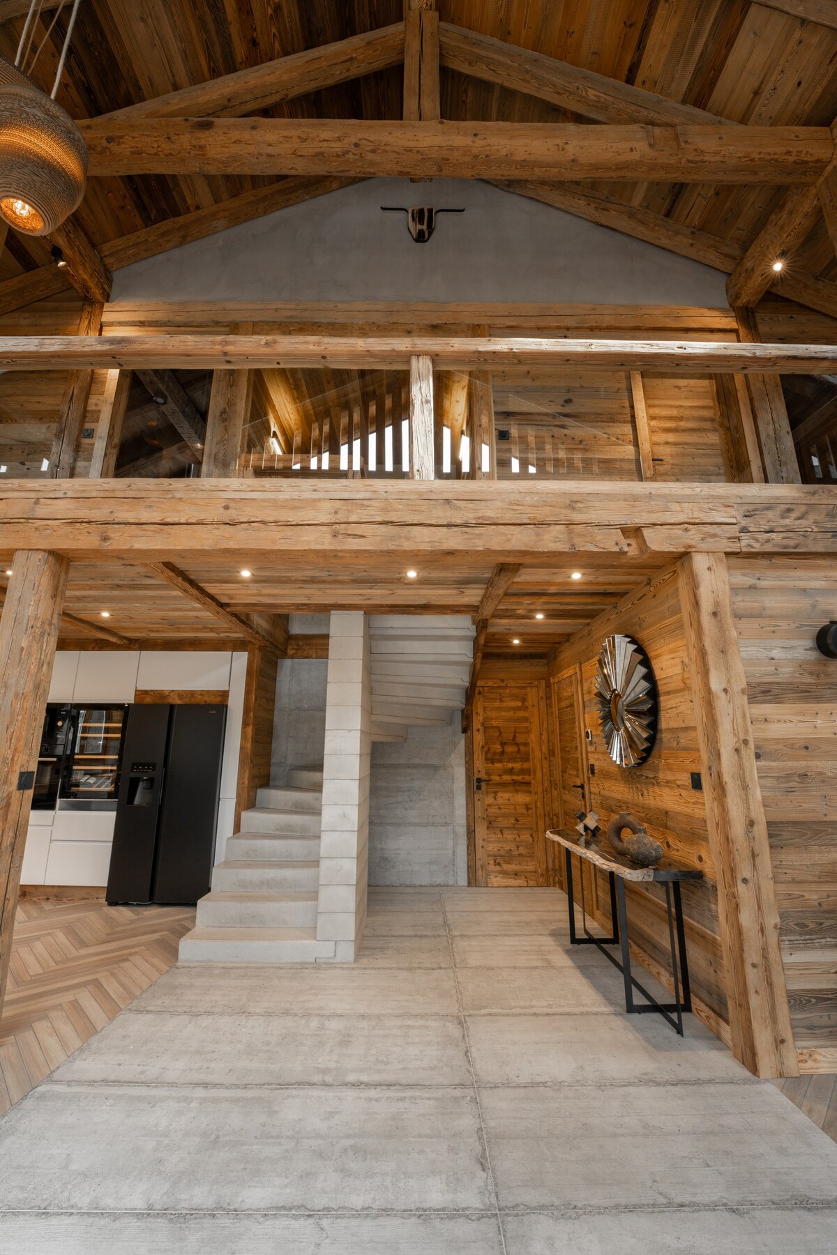 The interior features an elegant wooden design with high ceilings and exposed beams. A spacious staircase leads to an upper level, while a sleek, modern kitchen is visible in the background. The floor is a combination of polished concrete and patterned wood.
