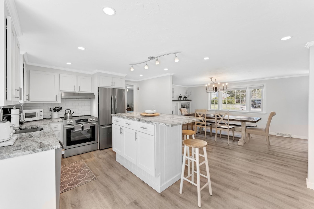 A modern kitchen is equipped with stainless steel appliances and ample counter space, featuring a spacious island with seating. An open dining area showcases a large table surrounded by chairs, illuminated by overhead lighting, creating a bright and welcoming space for gatherings.