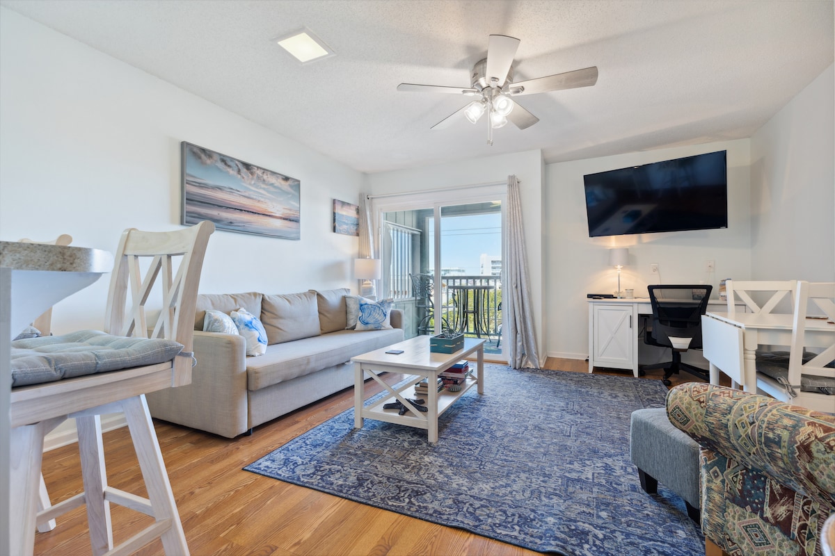 A light-filled living area features a comfortable sofa and a patterned armchair, positioned near a coffee table. A television mounted on the wall is complemented by a window with access to a balcony. Soft blue accents and a large area rug enhance the space.