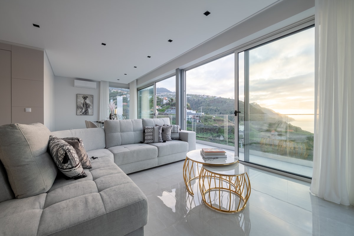 A spacious living area features a large sofa with decorative cushions, positioned near floor-to-ceiling glass doors. Natural light filters in, offering views of the surrounding hills and ocean. A circular coffee table with a gold base is centrally placed on the sleek tiled floor.