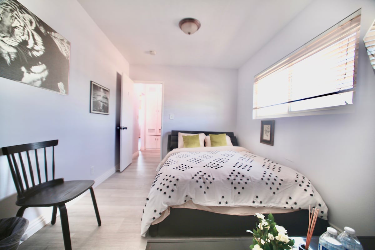 A cozy bedroom features a spacious bed adorned with a dotted blanket and decorative cushions. Natural light enters through a window with blinds. A black chair is placed nearby, and a small table with water bottles and flowers is visible, adding a touch of freshness to the space.