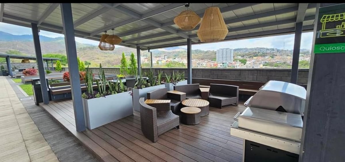 An outdoor social area features comfortable seating arrangements consisting of sectional sofas and circular tables. A barbecue grill is positioned nearby, and the space is adorned with plants. A scenic view of the surrounding hills and cityscape is visible in the background.