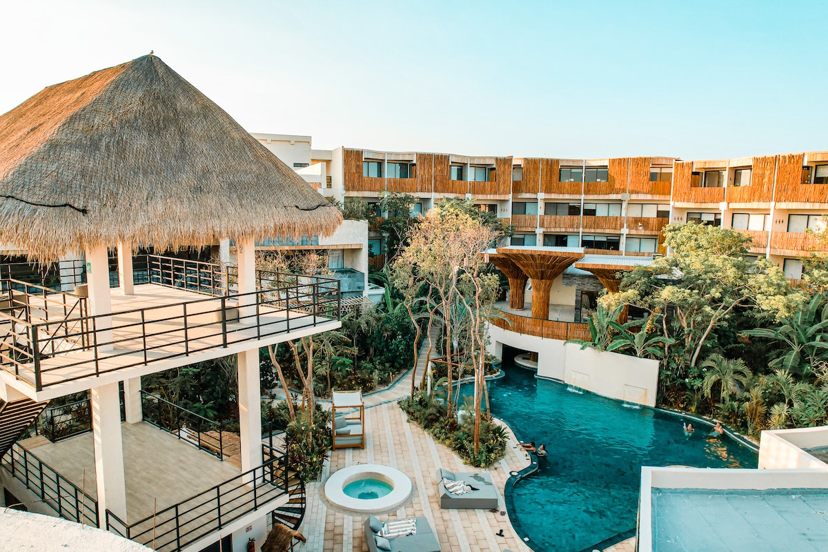 A multi-level resort area is visible, featuring a thatched-roof structure overlooking a lush courtyard. A large swimming pool surrounded by tropical plants is present, along with a smaller hot tub. Comfortable lounge chairs are arranged along the poolside, creating a relaxing outdoor atmosphere.