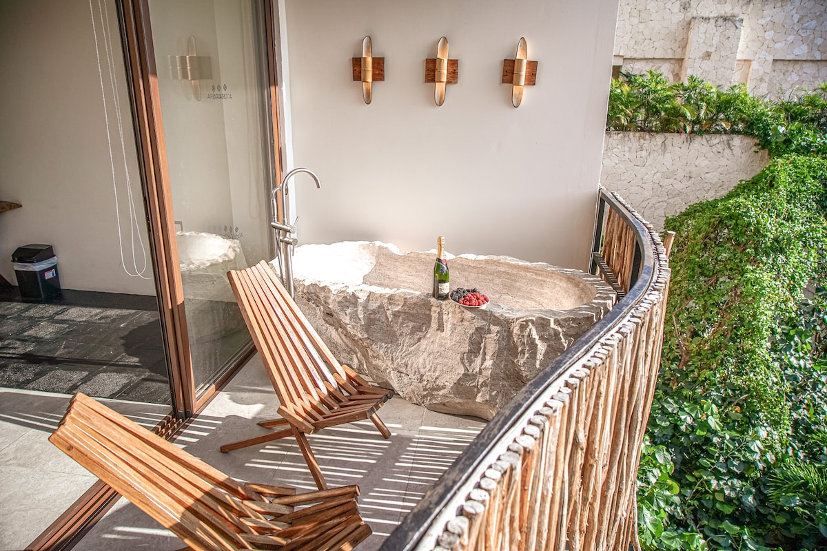 A private balcony features two wooden lounge chairs positioned next to a unique stone table. A bottle of champagne and fresh strawberries are arranged on the table. The space is surrounded by greenery, and glass doors provide access to the interior.
