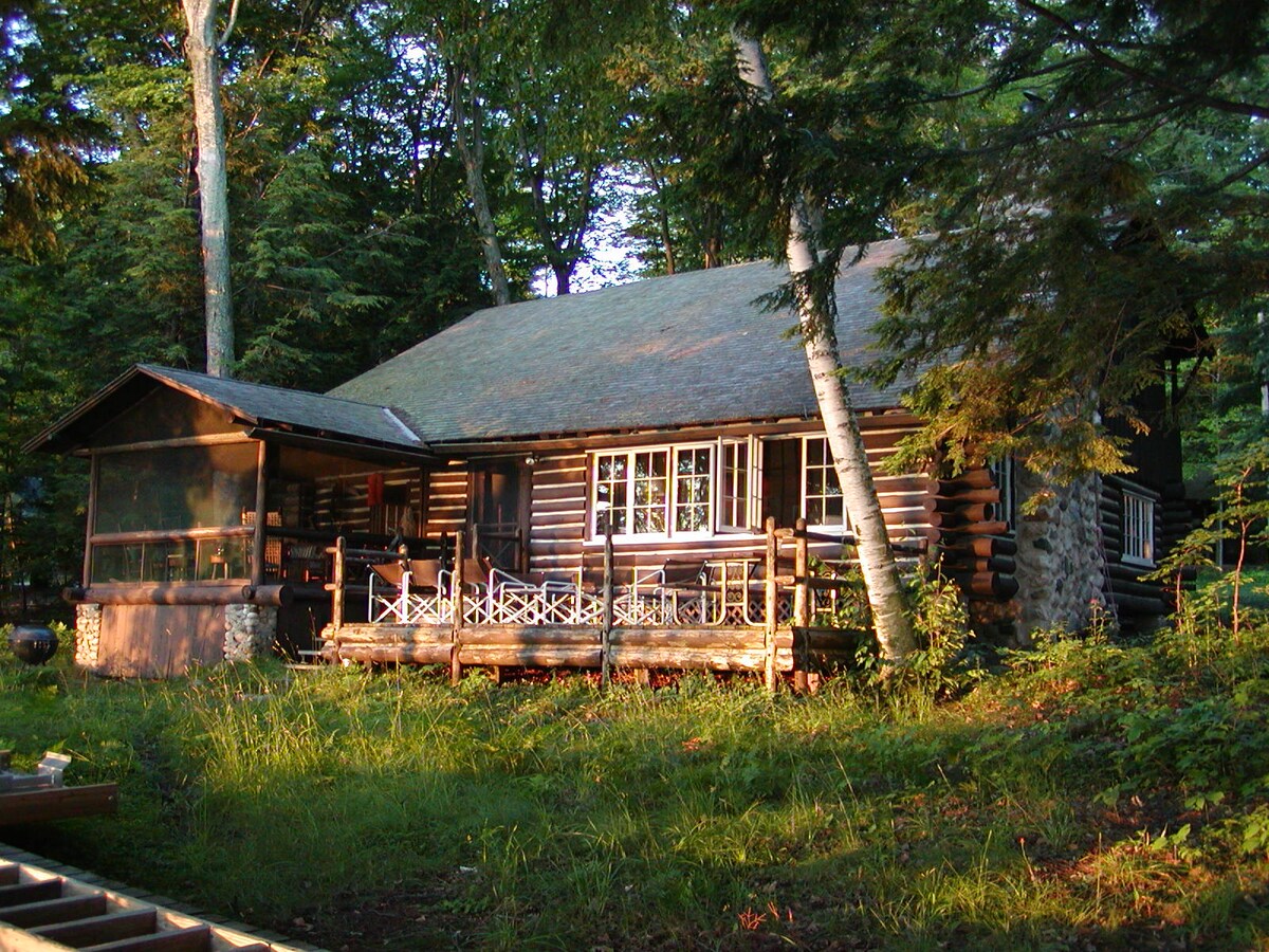 A log cabin is nestled among trees, featuring a welcoming porch with white chairs and a stone fireplace. The exterior showcases natural wood logs and a sloped roof, while a screened porch provides an additional outdoor space, inviting relaxation in a serene setting.