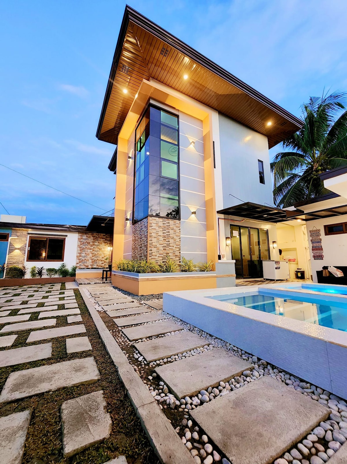 A modern two-story building is highlighted with large glass panels and a wooden overhang. Lush greenery surrounds the property, and a clear swimming pool reflects the evening sky. A neatly arranged stone pathway leads towards the entrance, contributing to the inviting outdoor space.