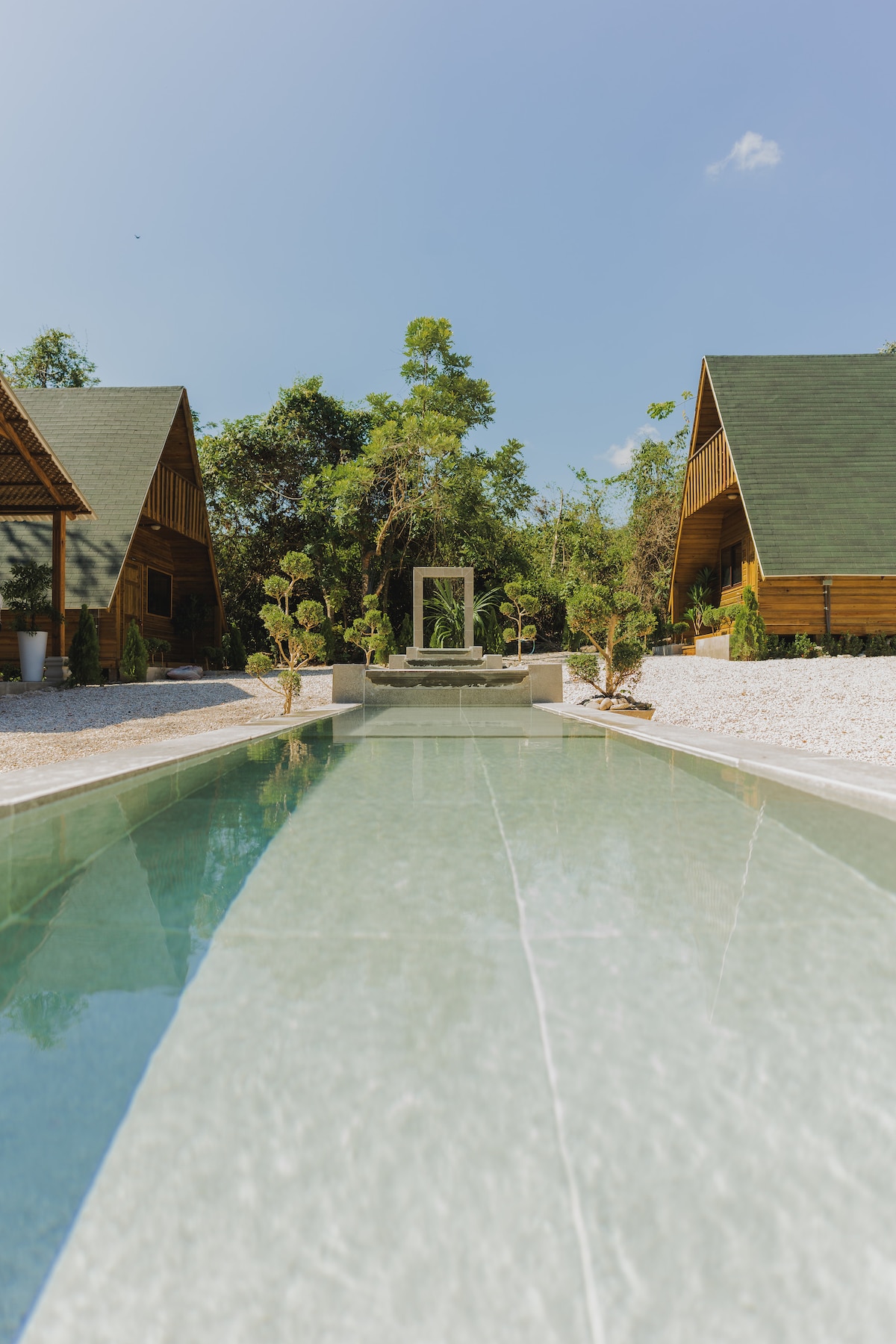 A clear swimming pool runs through the center of the property, bordered by white stone pathways. Two wooden cabanas with sloped roofs are visible in the background, surrounded by lush greenery and trees, offering a serene atmosphere.