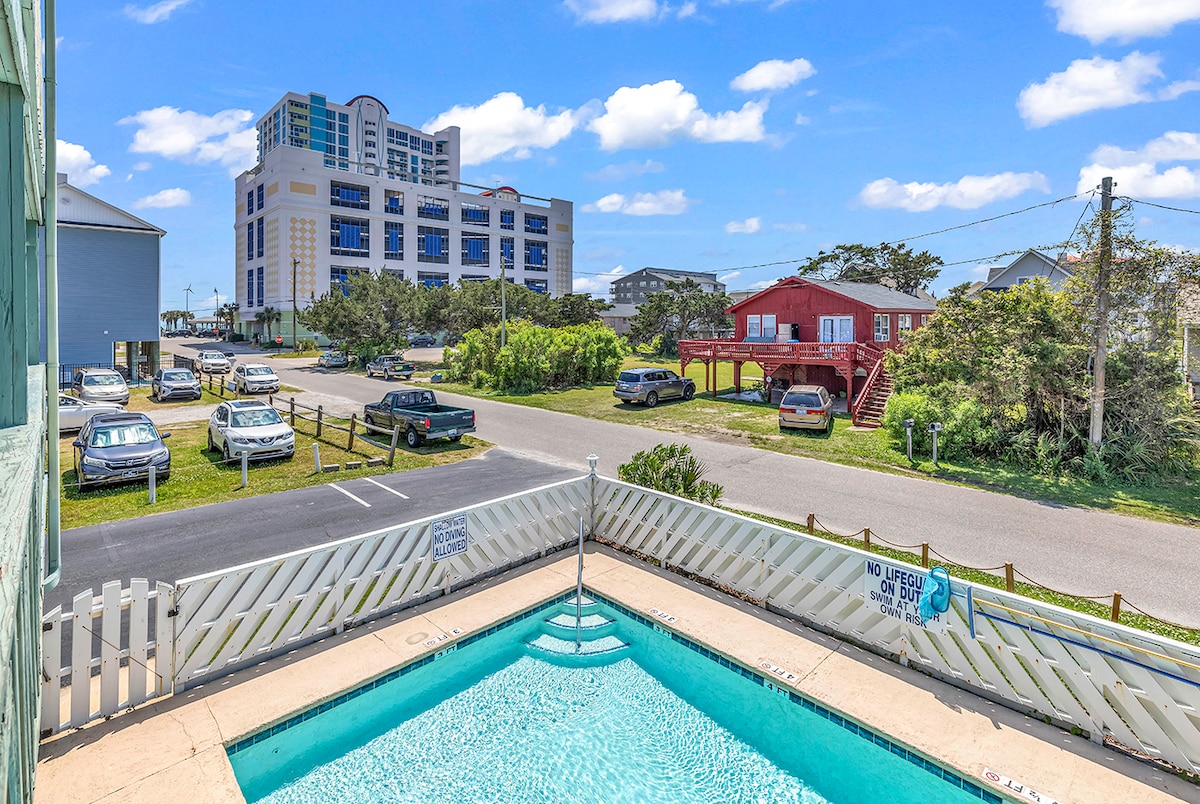 The image captures a view from a balcony overlooking a pool area. Various vehicles are parked nearby along a road lined with greenery. In the background, multiple buildings are visible, offering a sense of the surrounding area.