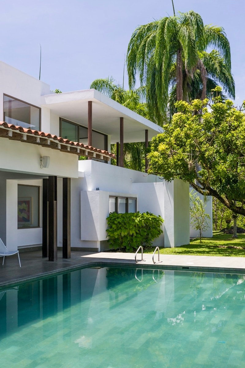 A modern villa is depicted, featuring a private pool surrounded by lush greenery. The exterior showcases a blend of white walls and wooden accents, with balconies overlooking the pool area. Tall palm trees add to the serene ambiance of the setting.