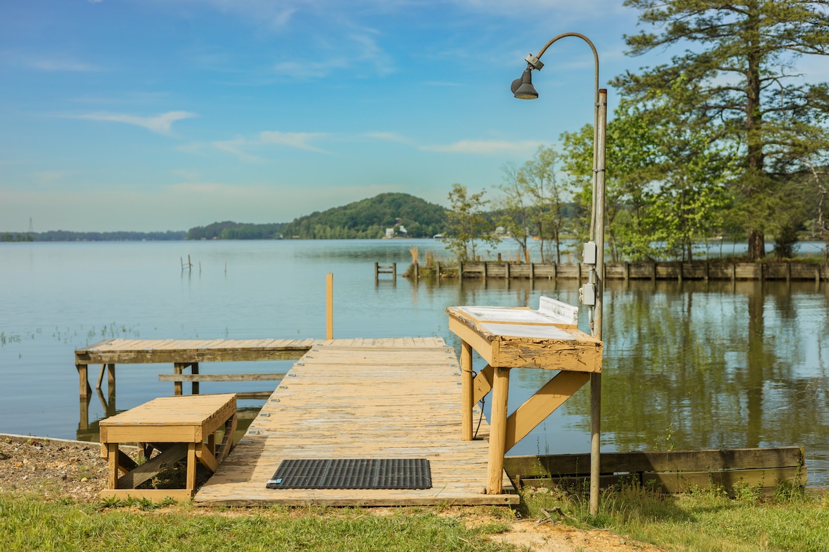 A wooden dock extends into Weiss Lake, featuring a fish cleaning station with a lamp overhead. The serene water reflects clear blue skies, surrounded by lush greenery. A small sitting area is present at the dock's edge, providing a peaceful spot to enjoy the views.