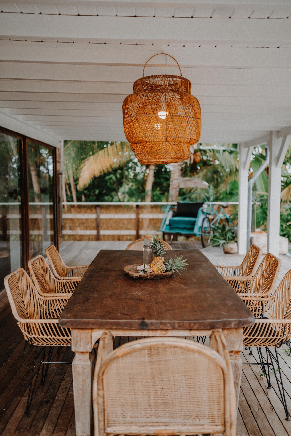 A spacious outdoor dining area features a natural wood table surrounded by six woven chairs. A decorative pendant light hangs above, creating a cozy atmosphere. A centerpiece of fresh pineapples is displayed on the table, complemented by lush greenery visible in the background.