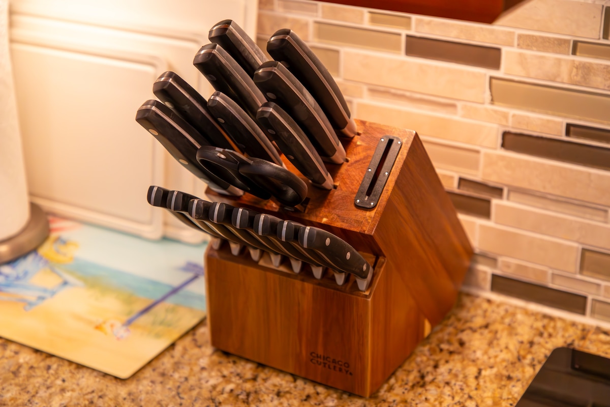 A wooden knife block sits on a granite countertop, holding a set of professional kitchen knives. The block features a modern design with multiple slots filled with various knife sizes, each with black handles. The kitchen background includes a light-colored backsplash and a glimpse of a paper towel holder.
