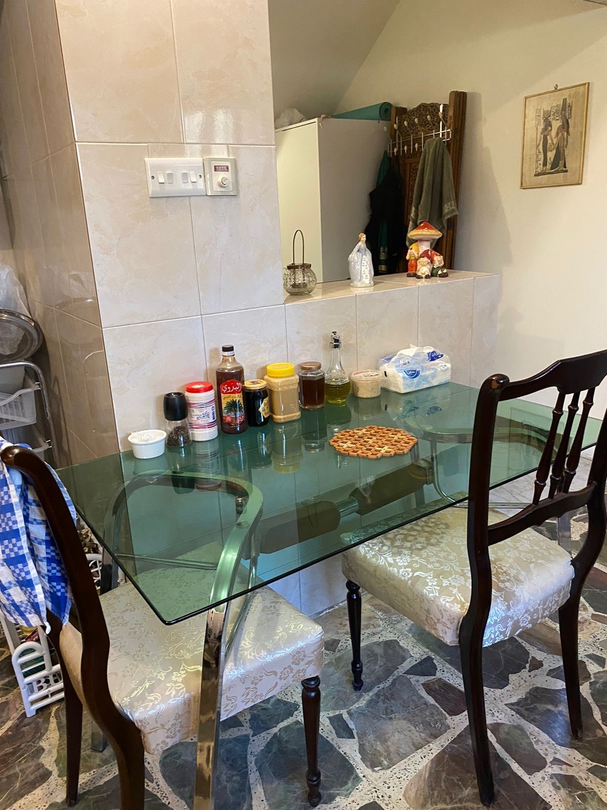 A glass dining table is set with two wooden chairs, each featuring cushioned seats. Condiments and snacks are arranged in the center, while a light-colored tiled wall serves as a backdrop. The space is illuminated with natural light.