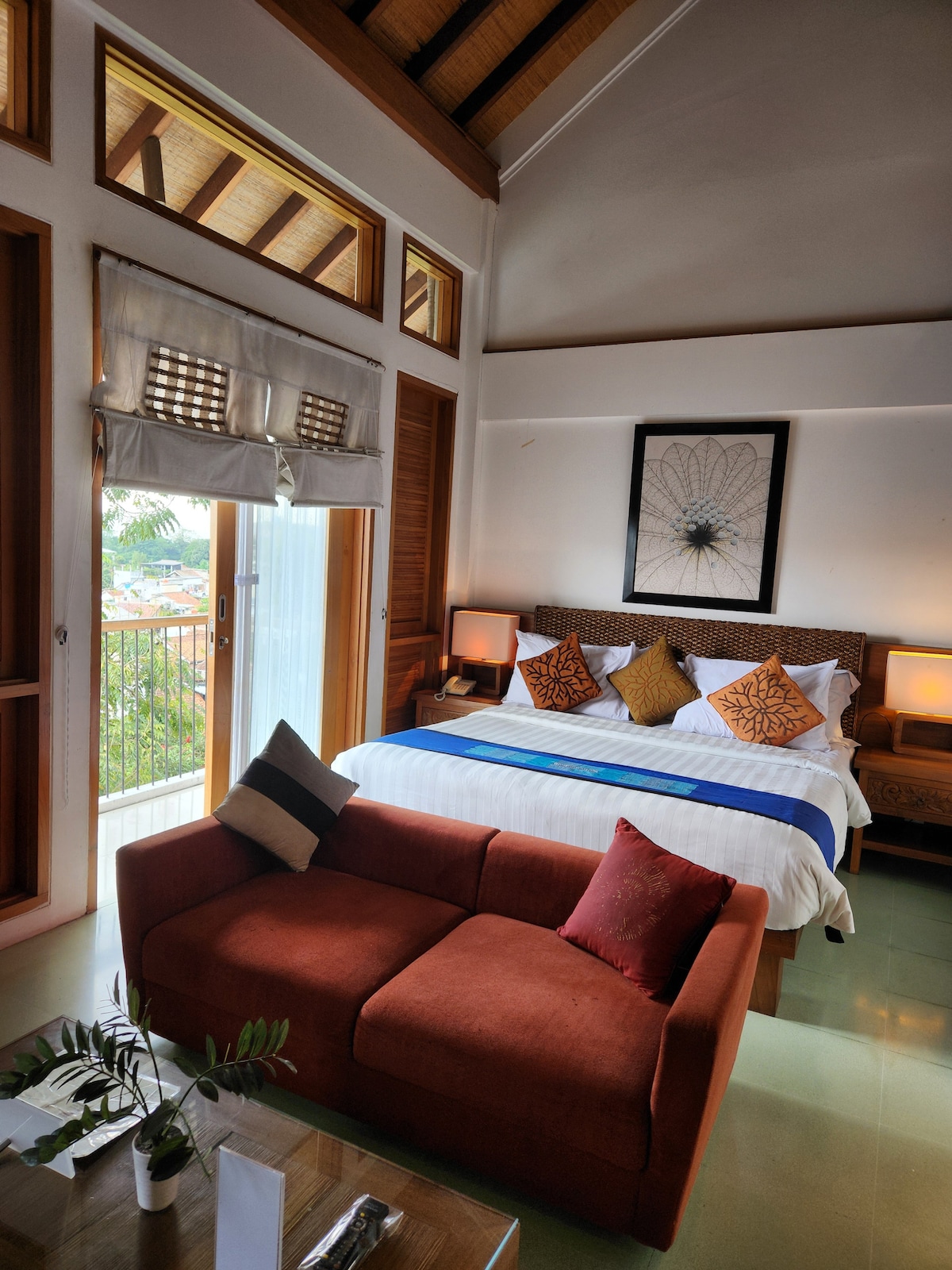 A bright and airy bedroom features a spacious bed adorned with decorative pillows. A small orange couch complements a coffee table, and natural light enters through large windows. A piece of wall art enhances the backdrop, providing a relaxing ambiance.