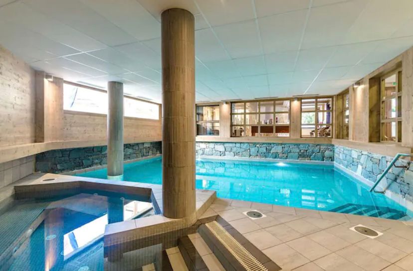 An indoor swimming pool is featured, surrounded by stone and wooden elements. Natural light enters through large windows, illuminating the sparkling water. Steps lead down into the pool, and a separate area with a seating space is visible in the background.