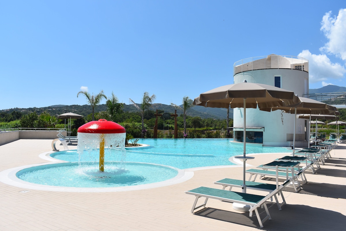 A spacious pool area features a vibrant red water fountain and lounge chairs arranged under umbrellas. Lush greenery surrounds the pool, and a modern building stands nearby, offering a serene environment for relaxation and leisure.