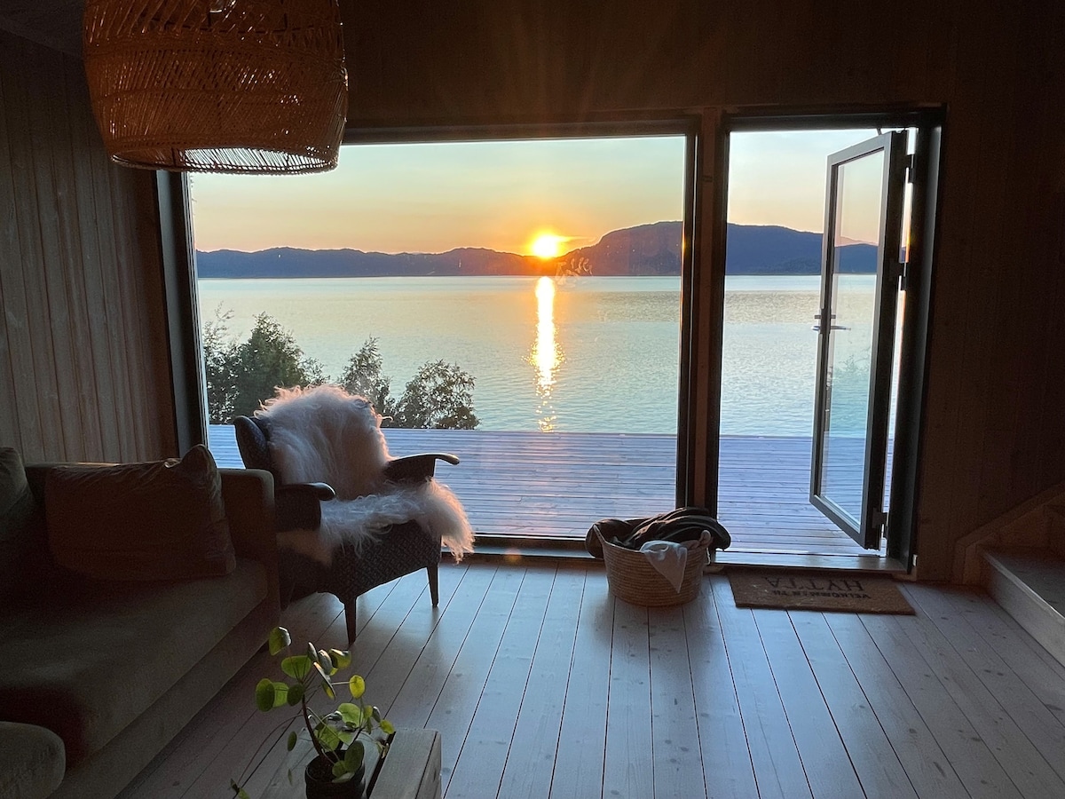A serene view is captured from the cozy interior, featuring an inviting seating area with a plush throw. Large glass doors open to reveal an expansive deck overlooking the tranquil waters of the fjord, illuminated by the warm glow of the setting sun.