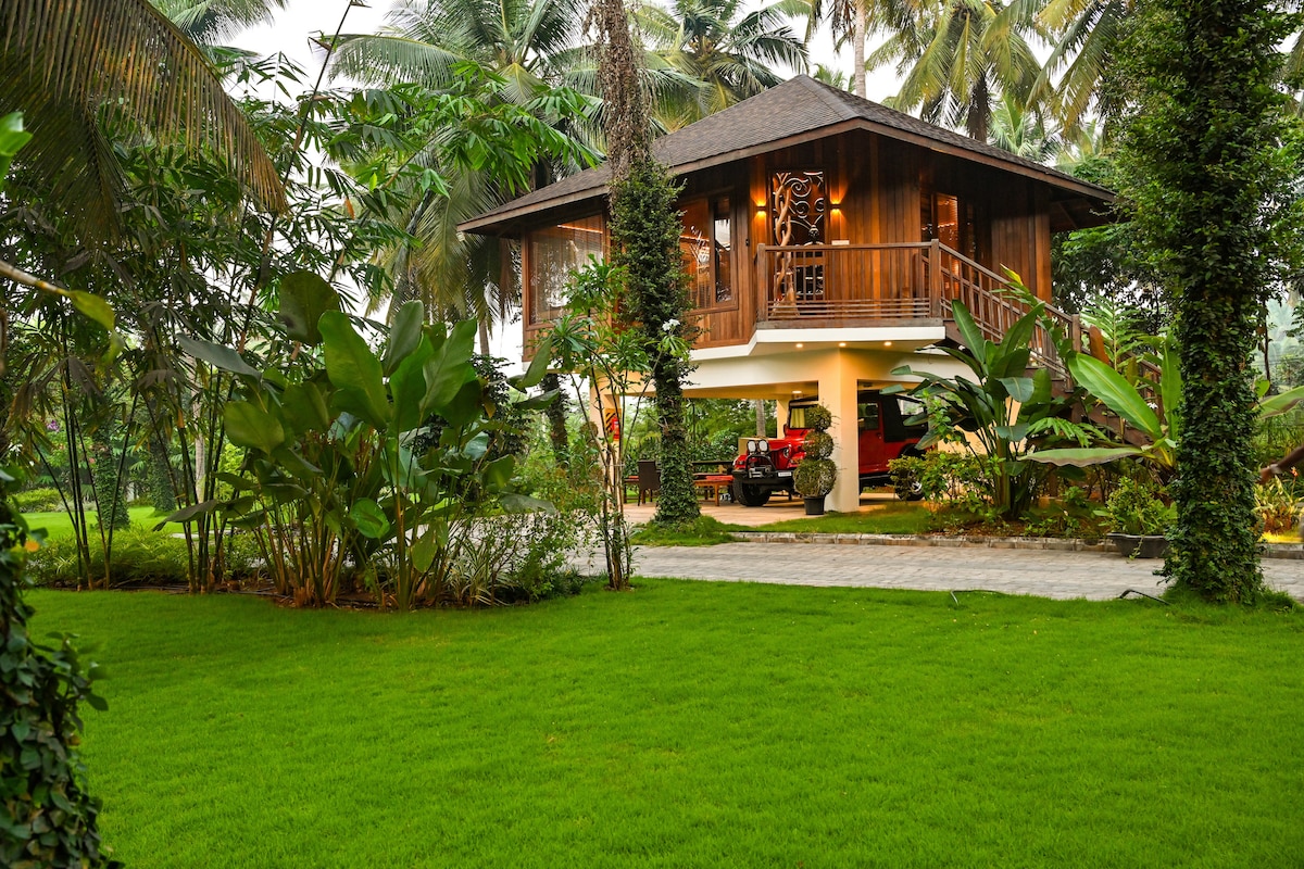 A wooden Balinese villa is showcased, elevated on stilts, surrounded by lush greenery. The tropical landscape features palm trees and vibrant plants, with a well-maintained lawn leading to the entrance. A pathway made of stone guides through the garden, enhancing the natural ambiance.