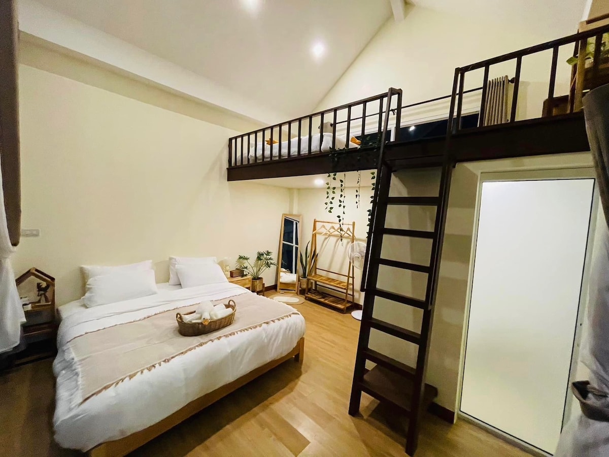 A bright room features a lofted sleeping area above a spacious bed with soft linens. Warm wood flooring complements the neutral wall tones. A cozy seating area is included, and natural light streams in from a nearby window, enhancing the overall inviting atmosphere.