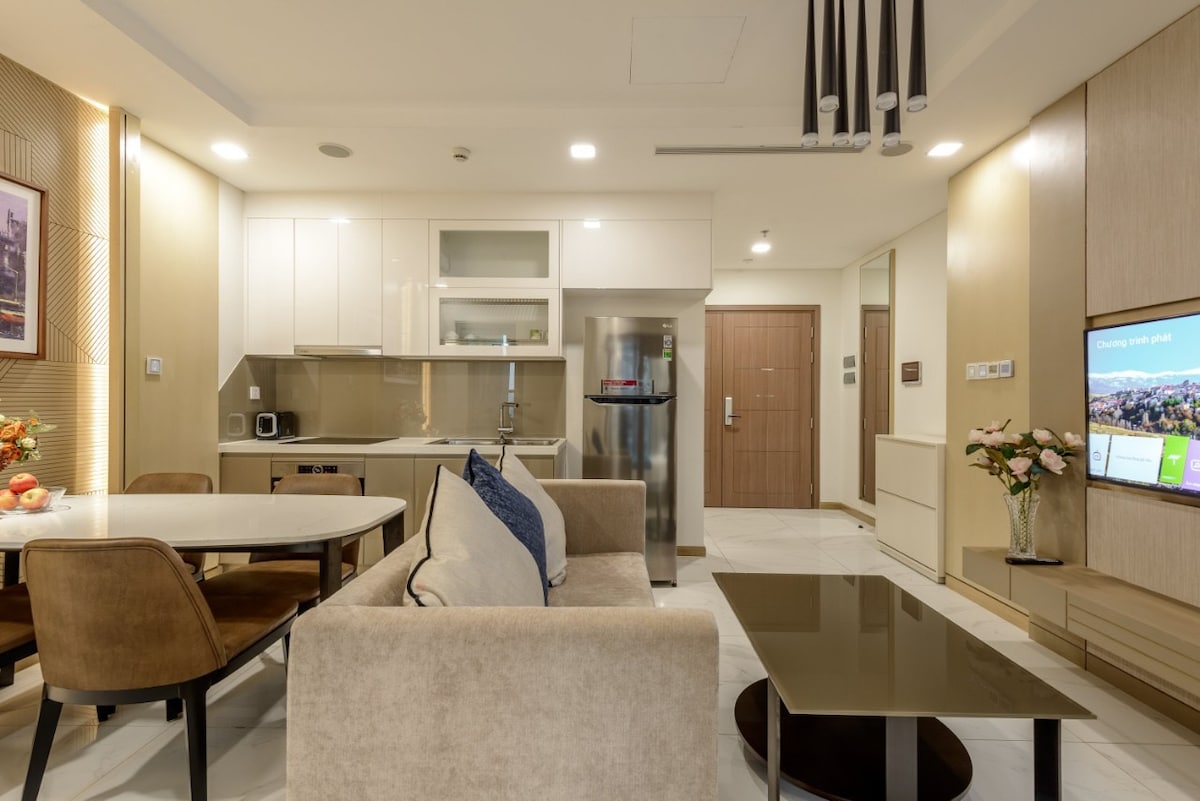 The living area features a neutral color palette with modern furnishings, including a comfortable sofa, a sleek coffee table, and a dining table with upholstered chairs. A smart TV is mounted on the wall, while a large window allows for natural light. The kitchen is equipped with contemporary appliances.