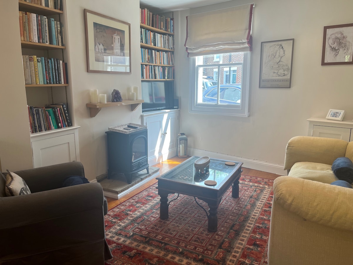 A cozy living room features a dark sofa and a light-colored armchair surrounding a glass-topped coffee table. Bookshelves filled with books line one wall. Natural light enters through a window, highlighting a decorative rug and a small wood stove underneath a shelf.