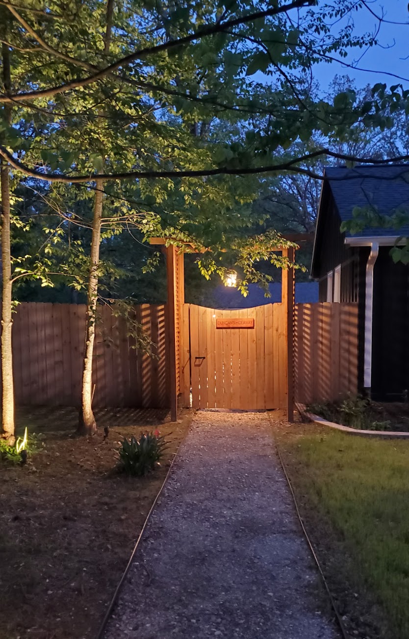 A serene pathway is illuminated by soft lighting, leading through a wooden gate surrounded by lush greenery. The gate, bordered by tall trees, creates a sense of privacy, inviting guests into a tranquil outdoor space beyond.