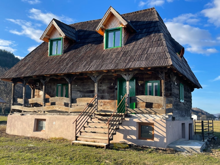 A charming wooden house is showcased, featuring a steeply pitched thatched roof and large green-framed windows. The porch, supported by sturdy wooden beams, is accessible via a set of steps. The scenic countryside provides a tranquil backdrop, with rolling hills in the distance.
