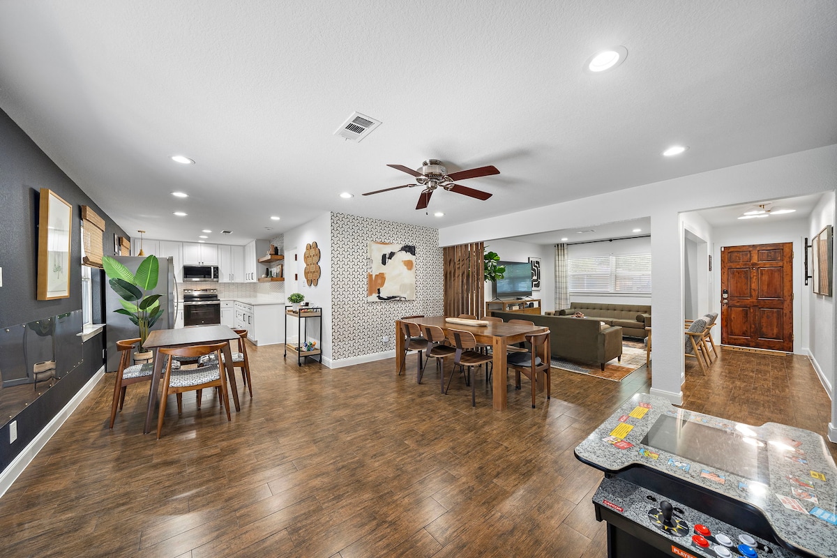An open concept living and dining area is highlighted by a spacious layout, featuring wooden flooring, a ceiling fan, and a mix of dining tables and chairs. Light gray walls create a modern backdrop for eclectic art pieces and large windows that provide ample natural light.